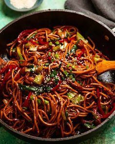 a wok filled with noodles and vegetables on top of a green tablecloth next to two spoons