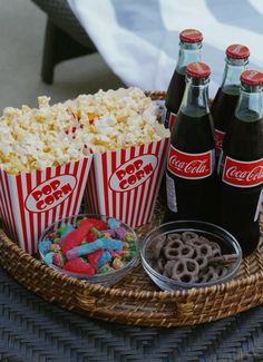 three sodas, popcorn and pretzels are sitting on a wicker tray