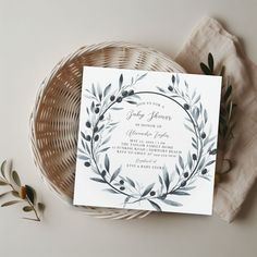 a wedding card with an olive wreath on it next to a wicker basket and flowers