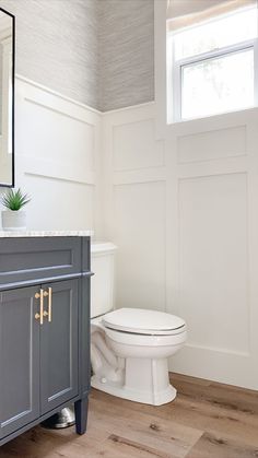 a white toilet sitting next to a gray vanity in a bathroom under a window with a potted plant on top of it