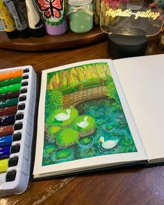 an open book sitting on top of a wooden table next to watercolors and markers