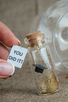 a person holding a tiny graduation cap in a glass bottle that says you did it