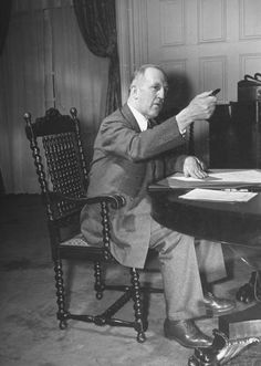 an old black and white photo of a man sitting at a desk pointing to something