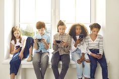 four children sitting on a window sill looking at their cell phones