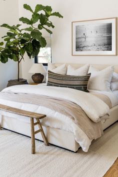 a bed with white sheets and pillows in a bedroom next to a potted plant