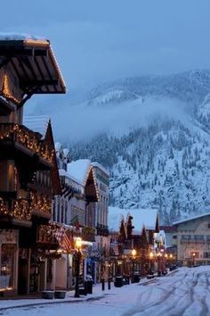 a snowy street lined with buildings and lights in front of a snow covered mountainside