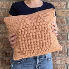 a woman holding a crocheted pillow in front of a brick wall with her hands on it