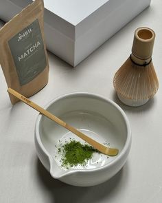 a white bowl filled with green stuff next to a bag of matcha powder and a wooden spoon