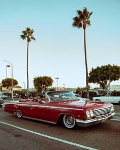 an old red car is driving down the street with palm trees in the back ground