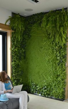 a woman sitting on a couch in front of a green wall with plants growing over it