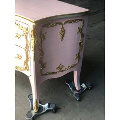 a pink and gold painted desk with ornate decorations on the top, next to two black casteors