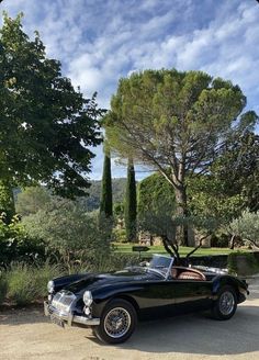 an old black sports car is parked in front of some trees and bushes on a sunny day