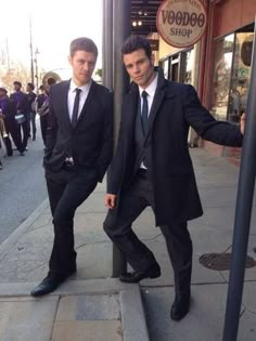 two men in suits are standing on the sidewalk near a sign that says voodoo shop