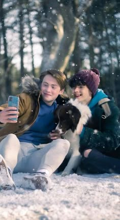 two women sitting in the snow with a dog and cell phone to their ear, taking a selfie