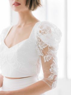 a woman wearing a white dress with sheer sleeves and lace on her arms, standing in front of a window