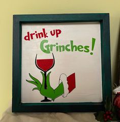 a framed sign that says drink up grinies with a wine glass in the foreground