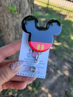 a person holding up a mickey mouse shaped id badge in front of a tree and fence