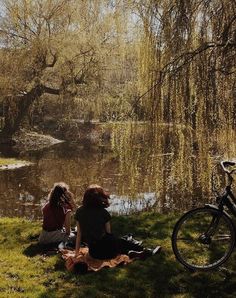 two women sitting next to each other on the grass near a river and bike in the background