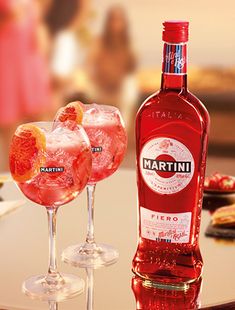 two glasses filled with pink liquid and garnished with orange peels sit on a table next to a bottle of martini