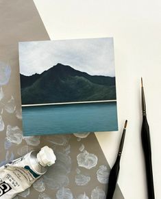 a painting and paintbrush on a table next to an empty card with mountains in the background