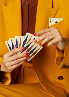 a woman in a yellow suit holding playing cards and wearing red nail polish on her nails