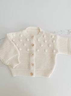 Soft and cozy, this stunning knit cardigan is the perfect addition to your little ones closet. Tiny Pom Pom appliqués all over. Textured pattern with front buttons. Cute Cotton Cable Knit Sweater, Playful Knit Long Sleeve Outerwear, Cute Cream Soft Knit Sweater, Playful Knit Cardigan For Winter, Cute Cream Cotton Cardigan, Playful Long Sleeve Knit Cardigan, Cute Long Sleeve Knit Cardigan, Playful Cream Winter Sweater, Cute Knitted Cotton Cardigan