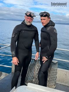 two men in wetsuits standing next to each other on a boat near the ocean