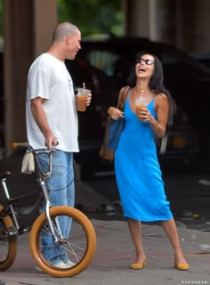 a woman in a blue dress standing next to a man on a bike and holding a drink