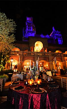 an outdoor dining area at night with lit up tables and chairs, in front of a castle