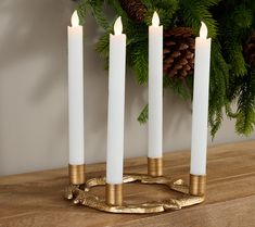 three white candles are sitting on a table next to evergreen branches and pine cones with gold accents