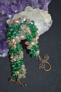 a green and gold beaded bracelet on a rock next to some purple crystal stones