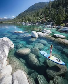 a woman is standing on a surfboard in the clear water near rocks and trees