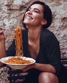 a woman sitting on a bench eating spaghetti from a white bowl with chopsticks