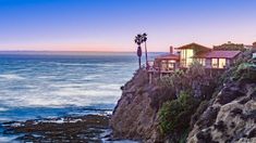 a house on the edge of a cliff overlooking the ocean at sunset with palm trees in the foreground