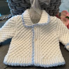 a crocheted white sweater with blue trim sits next to a pile of knitted blankets