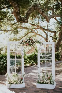 two white windows with flowers and greenery on the sides are set up for an outdoor ceremony