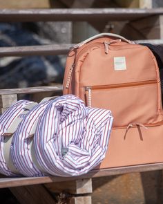 an orange backpack sitting on top of a wooden bench next to a pink bag and striped shirt