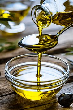 olive oil being poured into a glass bowl with olives in the background on a wooden table