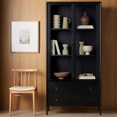 a black bookcase with glass doors and shelves in the corner next to a chair