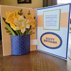 a birthday card with flowers in a blue vase on a wooden table next to a greeting card