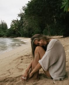 a woman sitting on top of a sandy beach next to the ocean with trees in the background