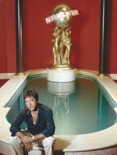 a man sitting in front of a fountain with a golden statue behind him on top of it