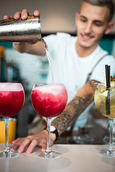 a man pouring drinks into two glasses at a bar with another one behind him and the other hand holding a shaker