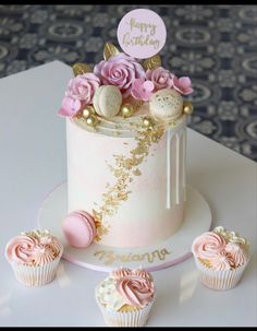 a white cake with pink frosting and gold decorations next to three cupcakes