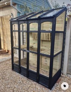 a small wooden greenhouse with glass doors on gravel ground next to fence and blue house