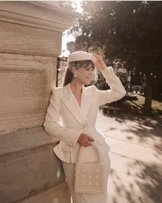 a woman leaning against a wall wearing a white suit and hat