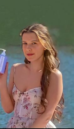 a woman in a dress is holding a bottle and looking at the camera while standing near water