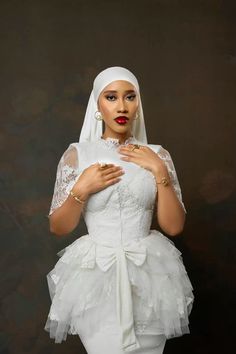 a woman wearing a white dress and veil with her hands on her chest, standing in front of a dark background