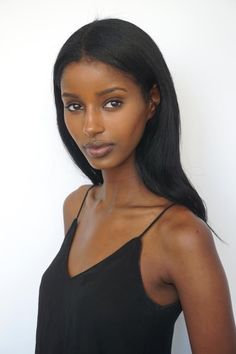 a woman with long black hair wearing a black top and looking at the camera while standing in front of a white wall