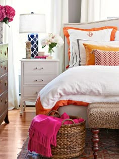 a bed with white sheets and orange pillows in a bedroom next to a dresser filled with flowers